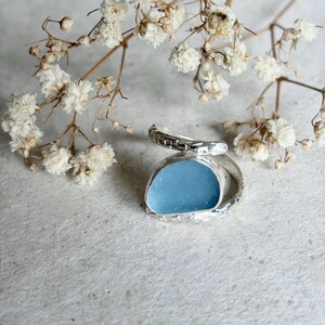 May include: A silver ring featuring a smooth, light blue, rounded piece of sea glass set within a textured, open-ended metal band. The ring is positioned near a sprig of small, dried white flowers with thin, branching stems.
