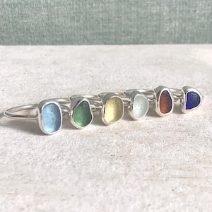 Peut inclure: Six bagues en argent, chacune ornée d'un morceau de verre de mer coloré. Les couleurs du verre de mer varient : bleu clair, vert, jaune, transparent, orange et bleu foncé. Les bagues sont présentées sur une surface claire.