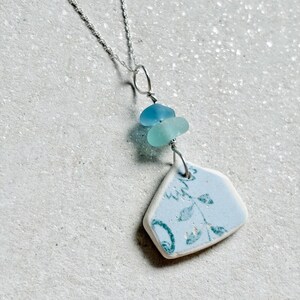 May include: A silver chain necklace featuring a pendant composed of two small, smooth sea glass beads in shades of blue and teal, suspended above a larger, flat, triangular piece of white ceramic with a light blue floral pattern.