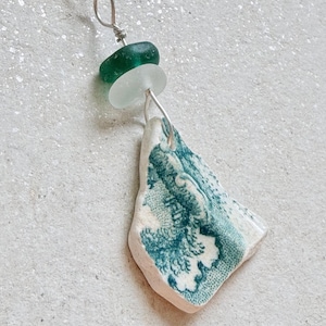 May include: A necklace featuring a silver chain with a pendant composed of a dark green sea glass bead, a frosted white sea glass bead, and a triangular shard of white ceramic pottery decorated with a teal blue floral or landscape pattern.