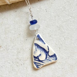 May include: A necklace featuring a silver chain and a pendant made from a triangular piece of white ceramic with a blue painted pattern. Above the ceramic piece are two small, rounded beads, one in translucent white and one in deep blue.