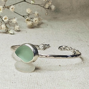 Könnte beinhalten: Ein silbernes Armband mit einem seegrünen Seeglasstein. Das Armband hat ein gehämmertes Band und einen Kettenverschluss. Das Seeglas ist in einer silbernen Lünette gefasst. Das Armband ist etwa 15 cm lang.