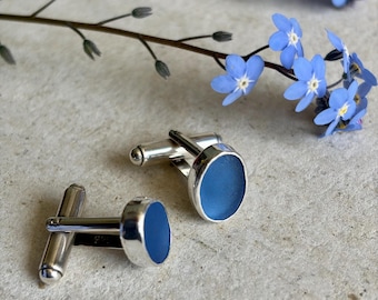 May include: A pair of silver cufflinks featuring smooth, oval-shaped matte blue inserts. The cufflinks have a polished metallic finish and a standard toggle closure mechanism, presented alongside small, delicate light blue forget-me-not flowers.