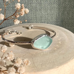 May include: A silver bangle bracelet featuring a hammered metal band and a single, smooth, light seafoam green sea glass stone set in a silver bezel. The bracelet includes a small silver chain extension with a circular clasp closure.