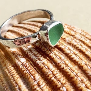 May include: A silver ring featuring a hammered band and a single, smooth, sea-glass-inspired green stone set in a bezel. The metal surface of the ring has a reflective, textured finish, and the green stone is shaped with rounded edges.