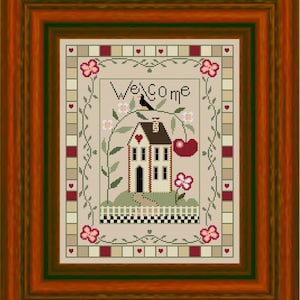 Può includere: Un ricamo a punto croce incorniciato che presenta una piccola casa con un cuore rosso sulla porta, un uccello appollaiato su un ramo e la parola "Benvenuto" in un font stilizzato. Il disegno è circondato da un bordo di cuori, fiori e viti.