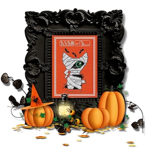 May include: A framed cross-stitch with a mummy cat design on an orange background, with the text "Mummy Cat". The black frame is ornate, with pumpkins, a lantern, and a witch's hat. Halloween-themed decoration.