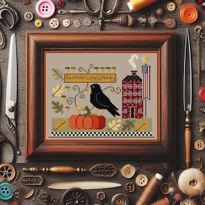 Può includere: Un ricamo incorniciato raffigurante un corvo nero posato su una zucca arancione. Il disegno include una casa rossa con una bandiera americana, il testo "Be Grateful Not Greedy" e foglie autunnali. Attrezzi da cucito circondano la cornice.