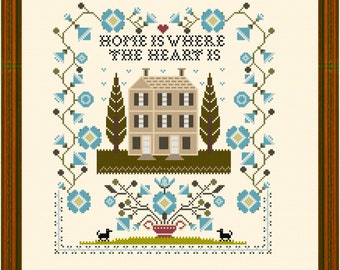 Puede incluir: Patrón de bordado de punto de cruz con una casa con un corazón encima y el texto "Home is where the heart is". El diseño está rodeado de flores azules y follaje verde. Dos perros negros están en la parte inferior del diseño.