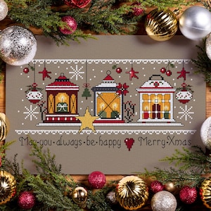 Può includere: Un ricamo a punto croce con tre lanterne festive rosse, bianche e dorate, con dettagli a stella e fiocco di neve. Il testo "May you always be happy Merry Xmas" è ricamato sotto. Il disegno è circondato da decorazioni natalizie e verde.