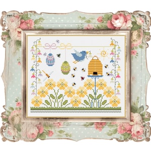 Può includere: Un ricamo a punto croce con un alveare, un uccello blu, fiori gialli e uova di Pasqua. Il disegno è incorniciato in una cornice bianca con un motivo floreale.