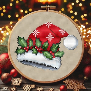 Può includere: Un disegno a punto croce di un cappello di Babbo Natale, incorniciato in un telaio da ricamo in legno. Il cappello è rosso con accenti di fiocchi di neve bianchi e un pompon bianco. Agrifoglio verde e bacche rosse adornano l'orlo, creando un tema festivo natalizio.