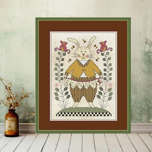 Può includere: Opera d'arte a punto croce incorniciata con un personaggio di coniglio stravagante. Il coniglio indossa una giacca gialla e pantaloni marroni, con la parola "Bunny" cucita in vita. L'opera è circondata da motivi floreali e di api, incorniciata in verde.