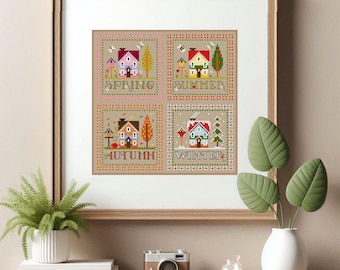 Op de afbeelding: Ingelijst kruissteekkunstwerk met vier seizoensgebonden scènes: Lente, Zomer, Herfst en Winter. Elke scène toont een huis, bomen en seizoensgebonden elementen. Het kunstwerk zit in een houten lijst en wordt tentoongesteld op een plank met een camera en planten.