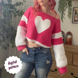 May include: A pink and white off-the-shoulder sweater featuring a large white heart. The sleeves have white stripes. The text "digital pattern" is on a white circle. The sweater is a cropped style.