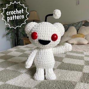May include: A white crocheted plush toy featuring a round head, two large red circular eyes, a black crescent-shaped mouth, and a black antenna with a white pom-pom on top. The text "crochet pattern" is visible in a white speech bubble.