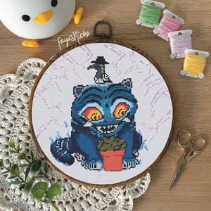 May include: A cross-stitch embroidery in a wooden hoop features a blue cat with a sinister grin, holding a potted plant. A small figure in a hat sits atop the cat's head. The background has a lightning bolt design. The text "fayesNiche" is visible.