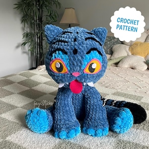 Può includere: Un gatto blu all'uncinetto con occhi gialli, lingua rosa e denti bianchi. Il gatto ha una coda a strisce nere e blu. Il testo "CROCHET PATTERN" è in un cerchio bianco.