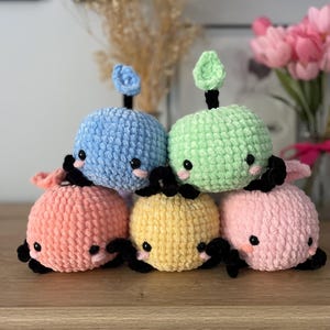 May include: Six crocheted plush toys shaped like whales in various pastel colors, including light blue, green, pink, yellow, and orange. Each whale has black eyes and a small crocheted fin on top.