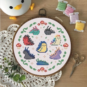 May include: A circular embroidery hoop with a white background and a green wreath design. The wreath is filled with colorful cartoon sea slugs and mushrooms. The text "fayesniche" is written in the upper right corner of the hoop.