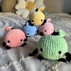 May include: Five crocheted plush toys in the shape of whales. The whales are in different colors: pink, light blue, yellow, green, and light pink. The whales have black eyes and mouths.