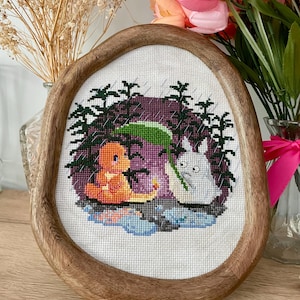 May include: A framed cross-stitch embroidery featuring two cartoon characters, a light orange creature and a white creature, in a green and purple forest setting. The embroidery is in a wooden oval frame.