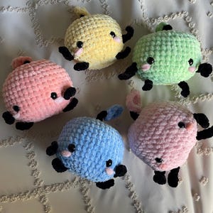 May include: Five crocheted plush toys in the shape of a cartoon animal. The toys are in various colors, including pink, blue, green, and yellow. Each toy has black eyes and a small mouth.