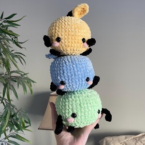 May include: Three crocheted stuffed toys stacked on top of each other. The top toy is yellow, the middle toy is blue, and the bottom toy is green. Each toy has black eyes and a small pink nose.