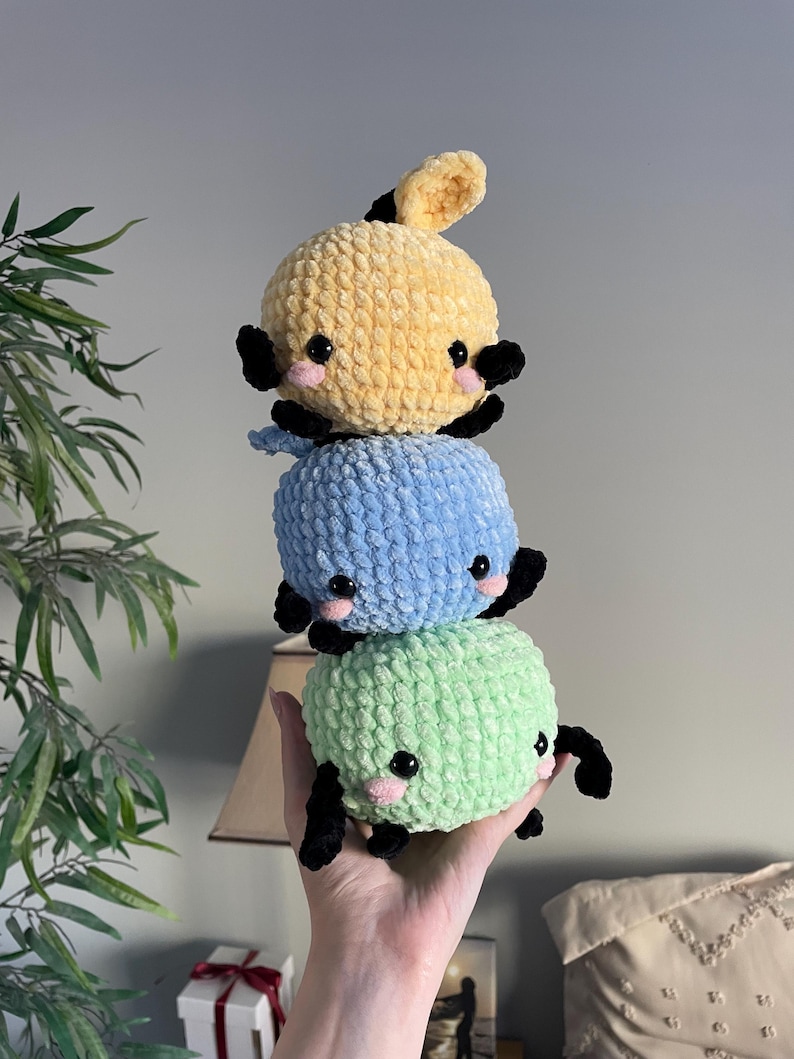 May include: Three crocheted stuffed toys stacked on top of each other. The top toy is yellow, the middle toy is blue, and the bottom toy is green. Each toy has black eyes and a small pink nose.