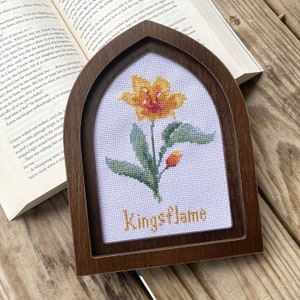 Puede incluir: Una bordadura de punto de cruz enmarcada que presenta una flor amarilla con hojas verdes y un capullo. La bordadura está sobre tela blanca y el marco es de madera marrón oscuro. El texto "Kingsflame" está bordado en amarillo debajo de la flor.