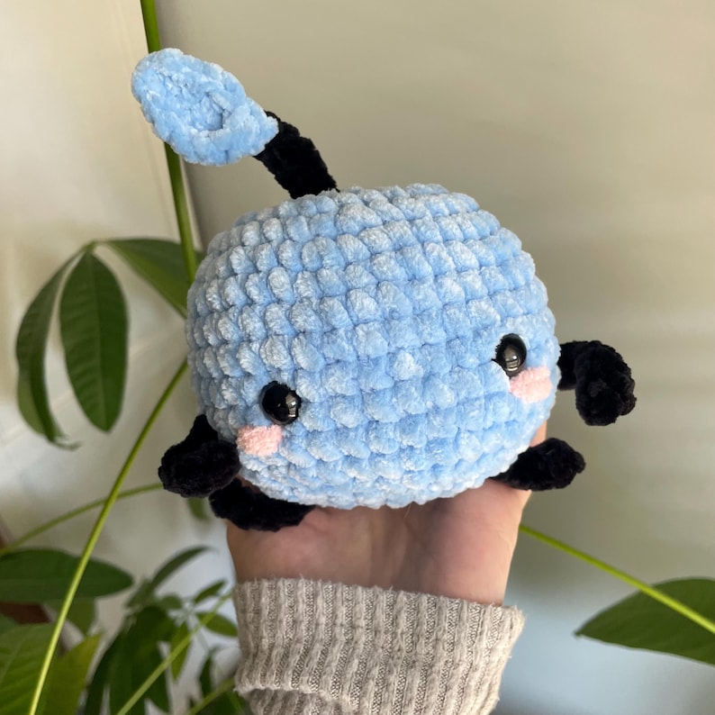 May include: A blue crocheted plush toy with black arms and a blue crocheted antenna. The toy has a cute face with black eyes and pink cheeks.