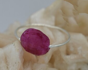 Puede incluir: Un anillo de plata con una piedra preciosa rosa en bruto. El anillo está colocado sobre una superficie blanca texturizada.