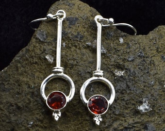 Könnte beinhalten: Ein Paar silberne Ohrringe mit roten Edelstein-Akzenten. Die Ohrringe haben ein kreisförmiges Design mit einem kleinen Edelstein in der Mitte.