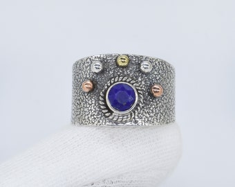 May include: A silver ring with a blue gemstone surrounded by a silver rope design. The ring is decorated with small gold and copper colored beads.