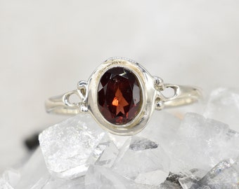 May include: A silver ring with an oval-shaped garnet gemstone. The ring has a delicate design with a simple band and a bezel setting for the stone.