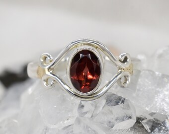 May include: A silver ring with a delicate, swirling design features a red oval gemstone in the center.