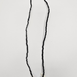 May include: A black cord necklace with a black arrowhead pendant, two white fang charms, and a silver clasp. The necklace is set against a white backdrop, highlighting its tribal design.