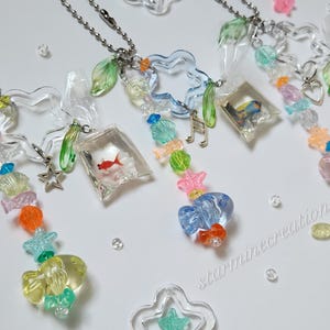 May include: Clear acrylic necklaces with colorful beads and charms. Each necklace features a chain, a fish-shaped charm, and a small clear bag with a goldfish inside. Other charms include stars, shells, and musical notes.