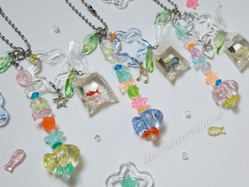 May include: Clear acrylic necklaces with colorful beads and charms. Each necklace features a chain, a fish-shaped charm, and a small clear bag with a goldfish inside. Other charms include stars, shells, and musical notes.