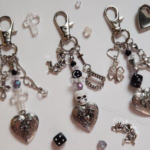 May include: Three silver keychains with charms. Each keychain features a silver locket with a heart design. The lockets are adorned with various charms, including a skull, a cross, a four-leaf clover, and a skeleton. The keychains are on a white background.