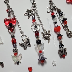 May include: Three silver charm necklaces with red, black, and white beads. Each necklace features a different charm, including a butterfly, a flower, and a fairy. The necklaces are strung on silver chains and have a safety pin clasp.