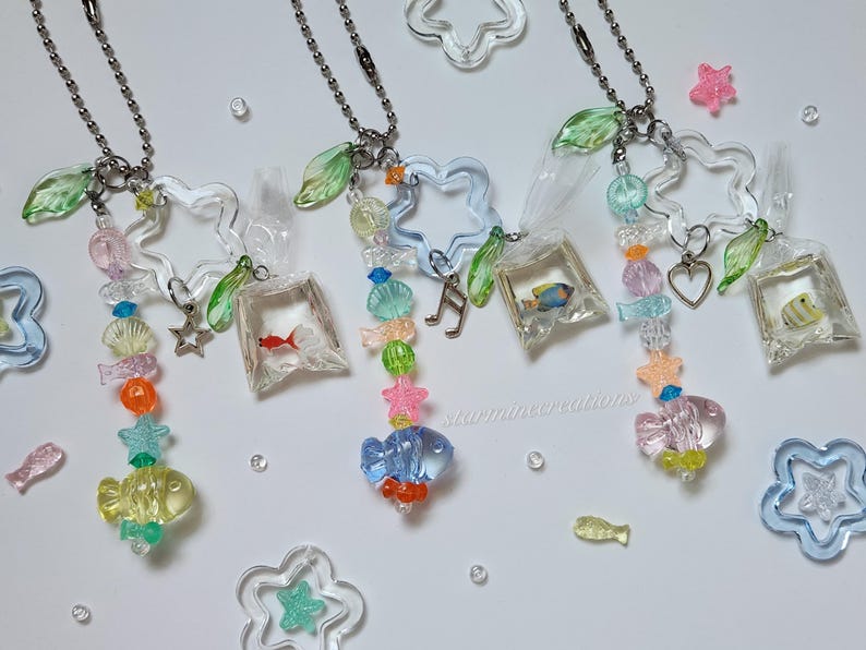 May include: Three clear acrylic fish-themed keychains with beaded accents. Each keychain features a fish-shaped charm, a small fish in a bag, and various colorful beads. The keychains are adorned with star and leaf charms.