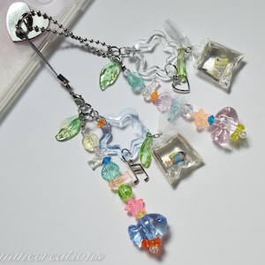 May include: A clear phone charm with a silver chain, featuring clear star and leaf-shaped beads, and small fish in plastic bags. Additional beads are in various colors, including blue, pink, and green. The charm is attached to a phone case.