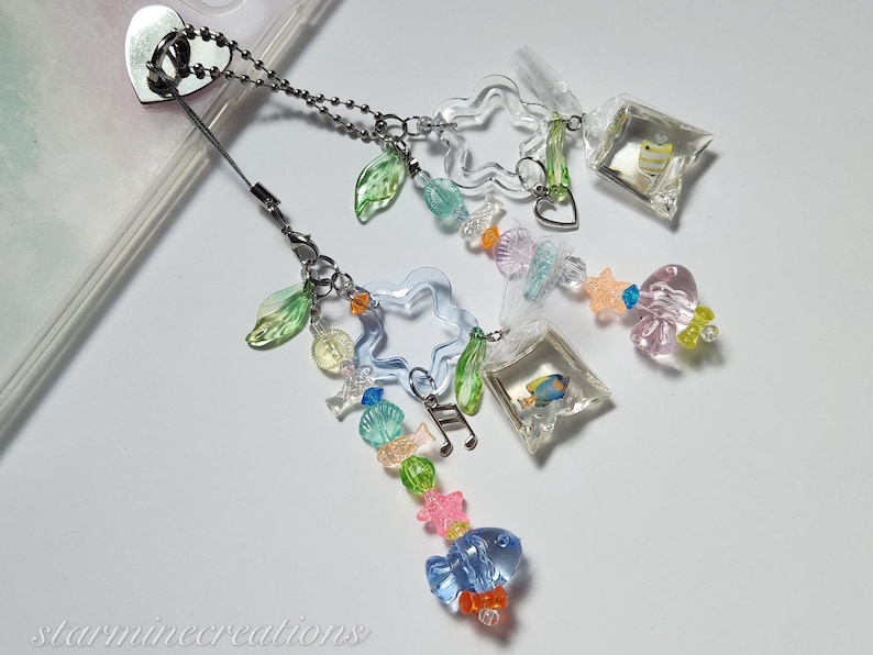 May include: A clear phone charm with a silver chain, featuring clear star and leaf-shaped beads, and small fish in plastic bags. Additional beads are in various colors, including blue, pink, and green. The charm is attached to a phone case.