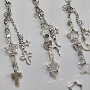 May include: Three silver charm keychains with star, cross, heart, and butterfly pendants. Each keychain features clear beads and a chain. The keychains are displayed on a white surface with scattered star-shaped charms.