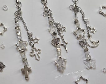 May include: Three silver charm keychains with star, cross, heart, and butterfly pendants. Each keychain features clear beads and a chain. The keychains are displayed on a white surface with scattered star-shaped charms.