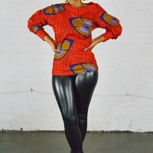 May include: A vibrant orange top with a geometric pattern in blue and yellow, paired with black leather leggings. The outfit is completed with leopard print heels, creating a bold and stylish look. The top features long sleeves.