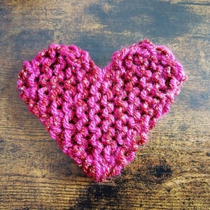 May include: A pink and red knitted heart with a sparkly finish. The heart is on a brown wooden surface.