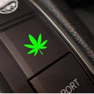 May include: Close-up of a black car interior button with a bright green cannabis leaf symbol. The leaf is centered on the button, contrasting against the dark background. The image suggests a custom car accessory.