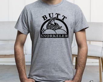 Op de afbeelding: Een sportgrijs t-shirt met korte mouwen en een zwarte grafische print op de borst. Het ontwerp toont een snorkelmasker boven twee ronde vormen, met de tekst BUTT SNORKELER in een boog boven en onder de centrale illustratie geplaatst.